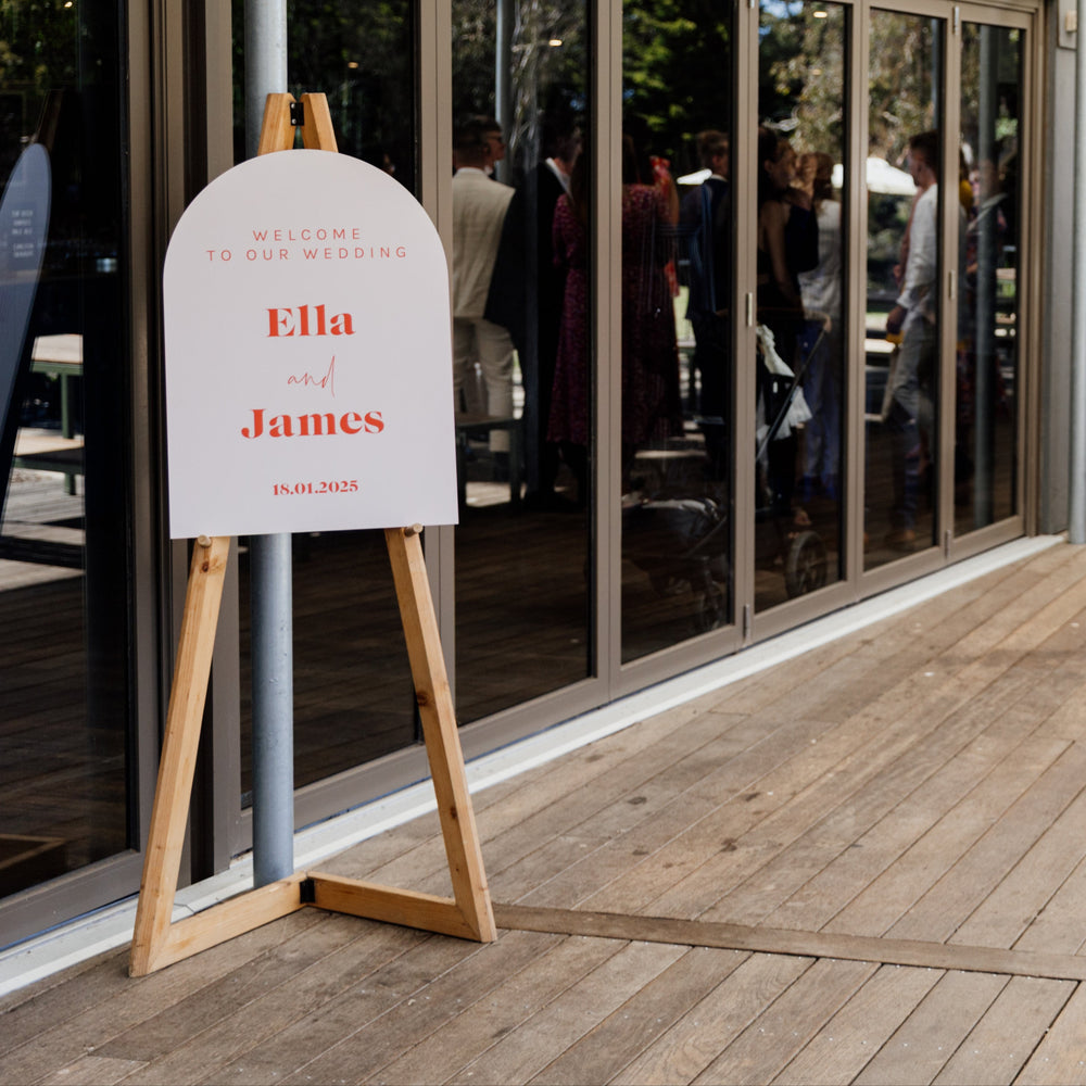 Easel with a sign reading 'Ella and James' in an outdoor setting with people and a building.