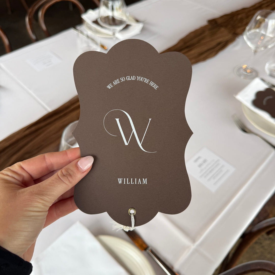 A hand holds a scalloped brown place card with a white monogram and the name WILLIAM, photographed over a decorated event table with white linens, glassware, and chairs.