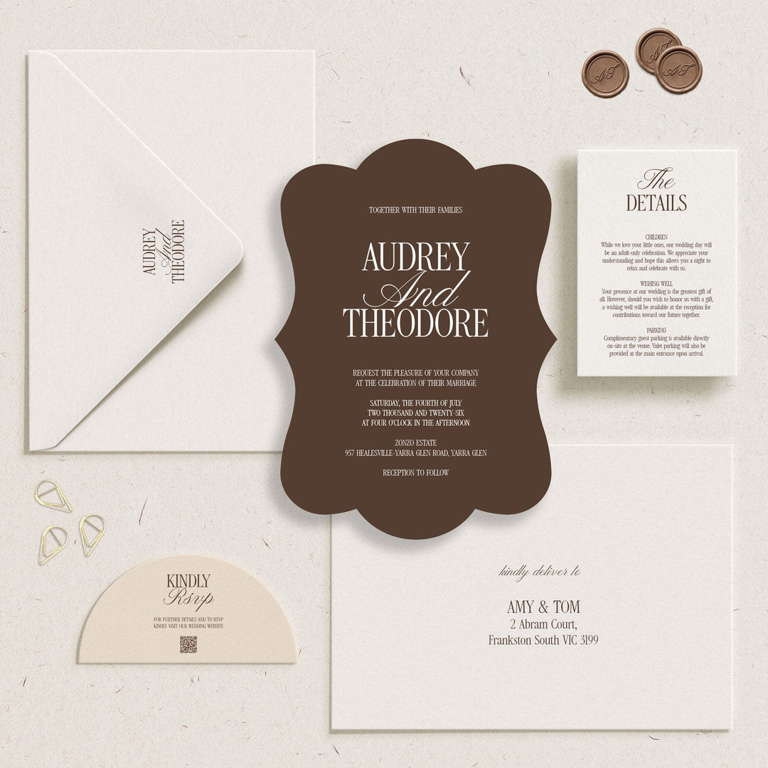 A wedding invitation suite laid out on a textured cream background, featuring a dark brown central invitation card, a white envelope, a white details card, a cream RSVP card, and three bronze coins as styling props.
