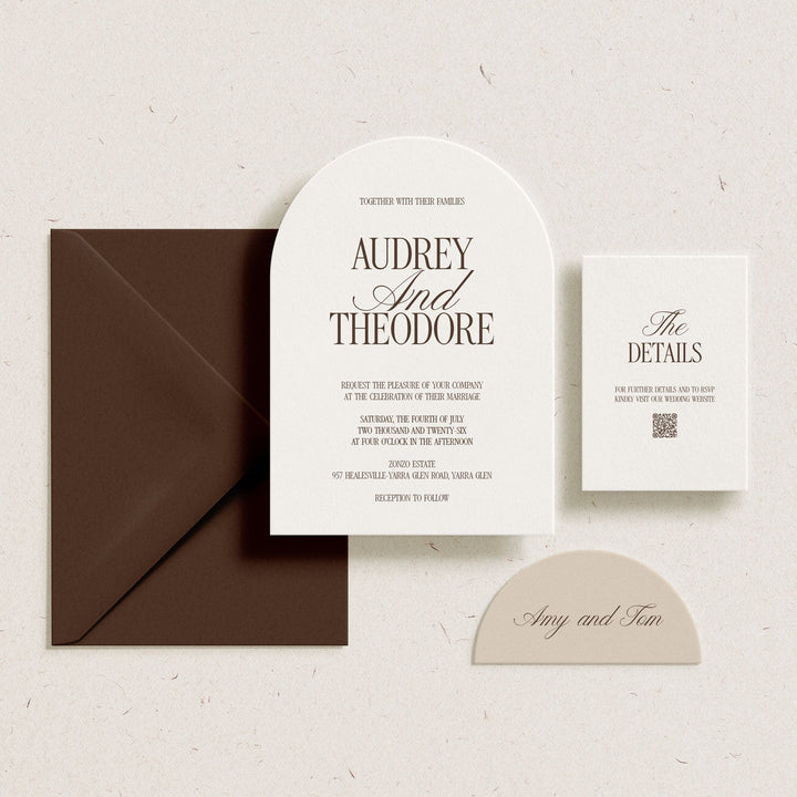 Wedding invitation suite on a textured off-white background, featuring an arched ivory invitation card, a dark brown envelope, a small details card, and a matching cream place card.