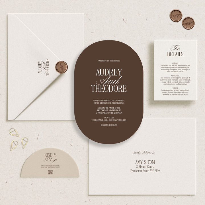 A coordinated wedding invitation suite laid out on a textured off-white surface, including a large oval brown invitation card, a white envelope with a wax seal, a details card, a small contact card, and a few decorative gold and brown wax seals.