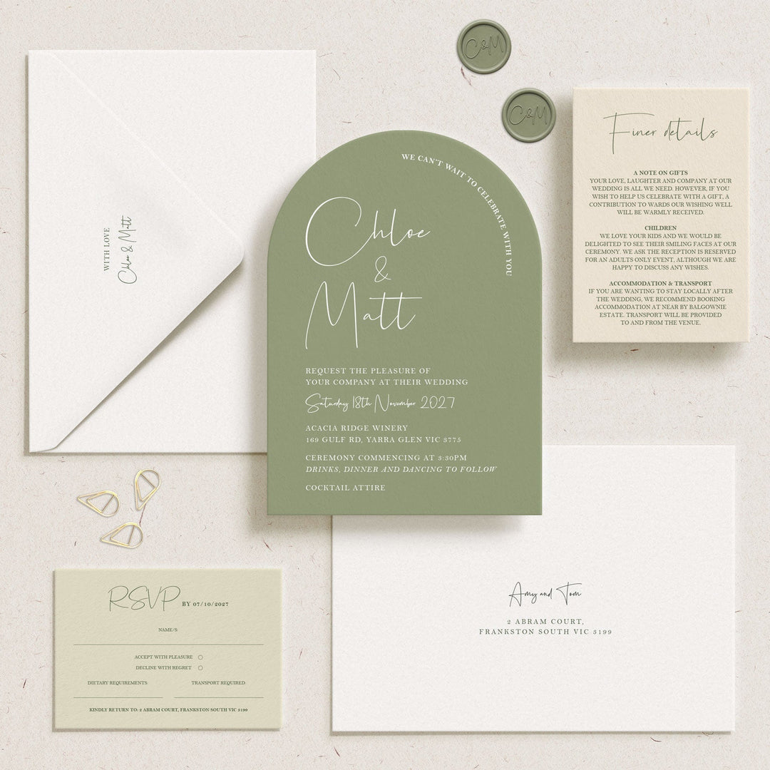 A coordinated wedding invitation suite laid out on a light textured surface, including a muted sage green arch-shaped invitation card, white envelopes, a cream details card, a pale green RSVP card, and two small metallic confetti pieces.