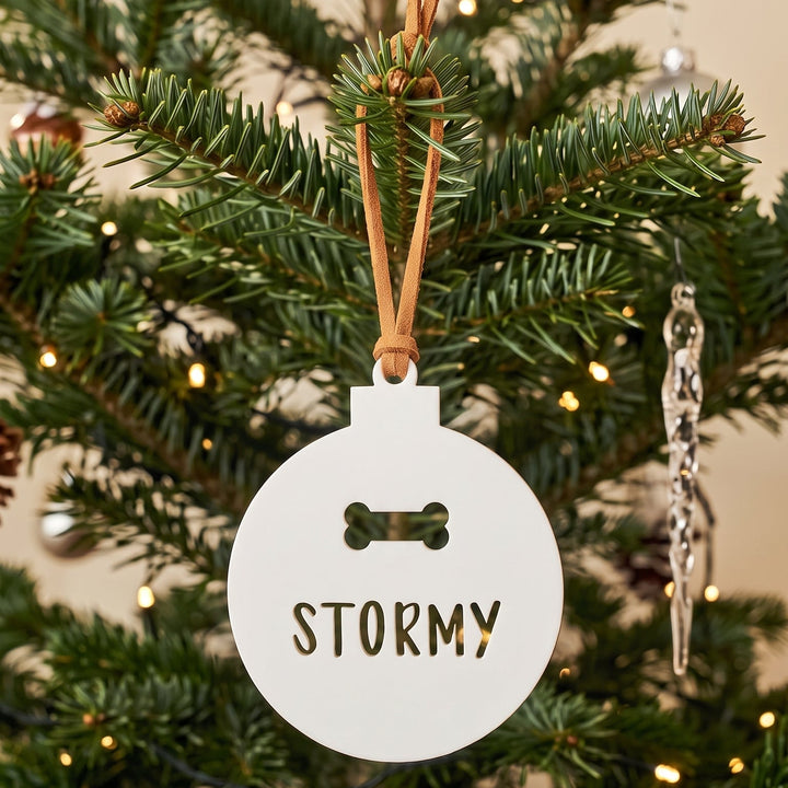 A round white Christmas ornament hanging on a pine tree, printed with a small bone icon and the name &