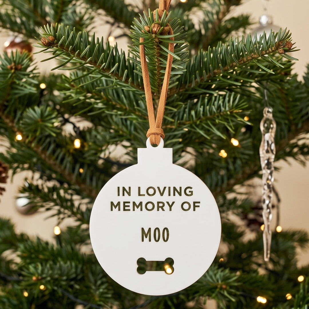 A round white pet memorial Christmas ornament hanging on a green Christmas tree. The ornament features the text &